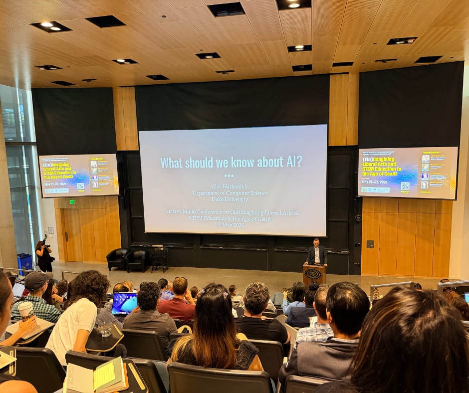 A large crowd of conference attendees fills a lecture hall, facing the front where Alex Hartemink's title slide reads "What should we know about AI?"