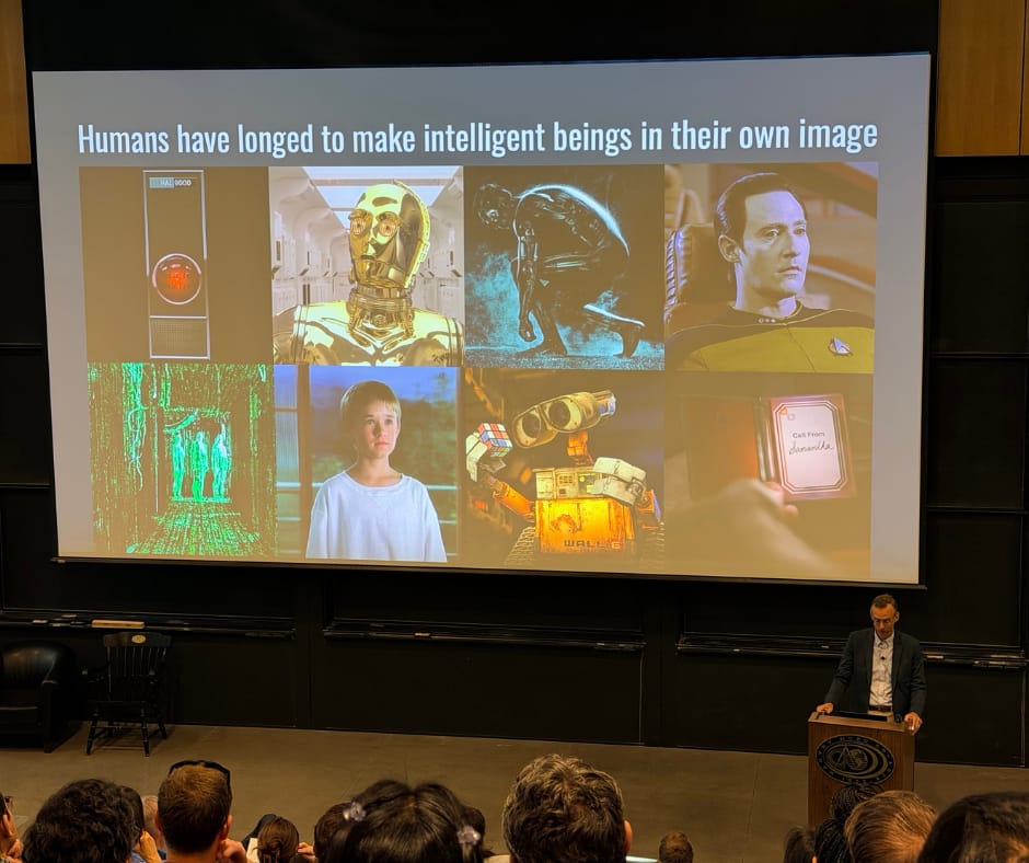 Alex Hartemink presenting from the front of a large lecture hall. His slide is titled "Humans have longed to make intelligent beings in their own image" and shows a collage of film stills, including HAL 9000, C-3PO from Star Wars, TRON, Data from Star Trek, a young boy in a white sweatshirt looking out a window toward mountains and trees, three shadowy green-iridescent figures in a room with glowing floors, walls, and ceiling, WALL-E holding a Rubik's cube, and a hand holding a foldable device showing a call from Samantha in the film Her.