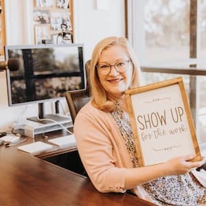 Dave & Bonni || Headshots Woman sits at a desk, holding a sign that reads: "Show up for the work."
