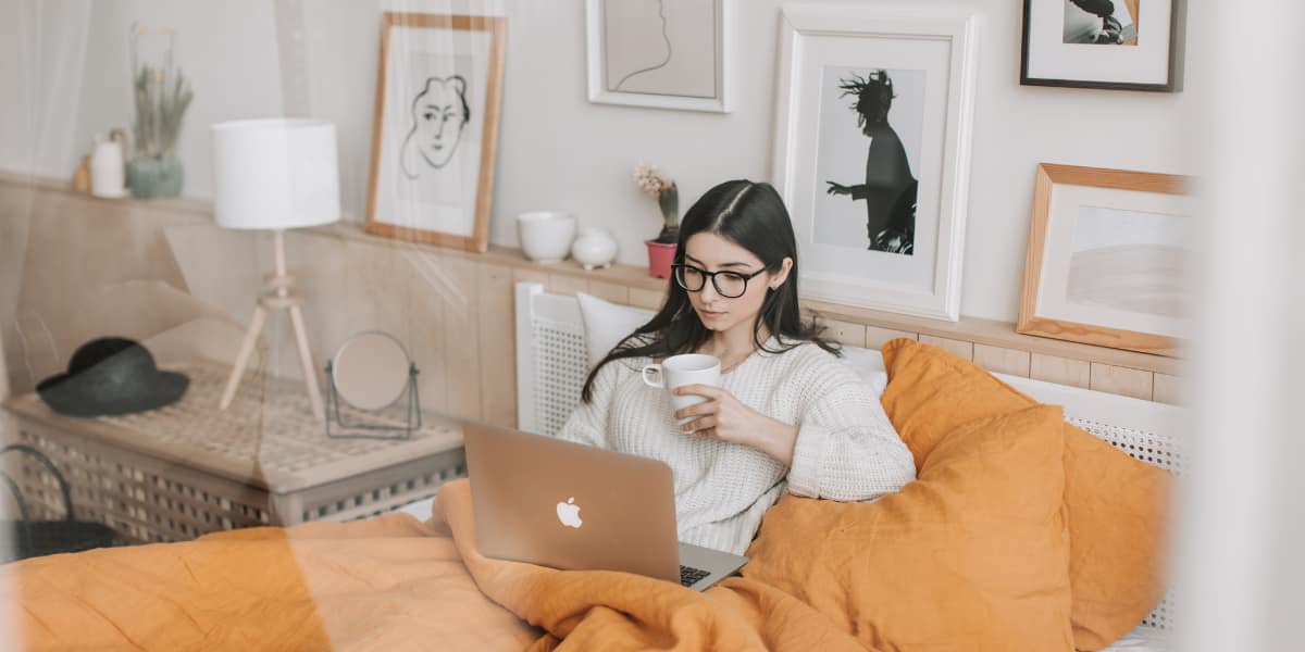 woman uses a Mac laptop in bed with a cup of coffee