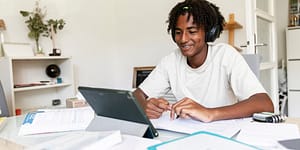 young man sits at a desk with headphones on looking at his laptop screen