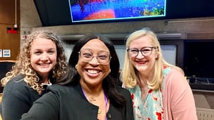Three women stand together with a brightly colored slide being projected in the background