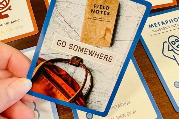 Person holding a card from a deck of cards, which says "Go Somewhere" on it and features a travel journal and bag.