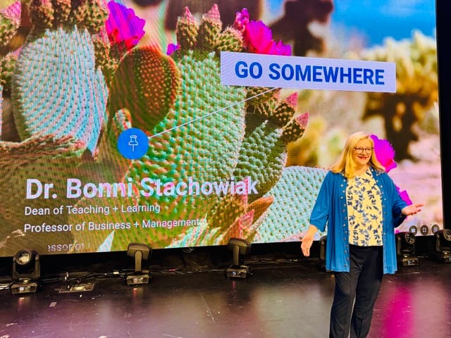 Woman stands in front of a colorful slide that says: "Go Somewhere" on it. Lots of desert themed flora...