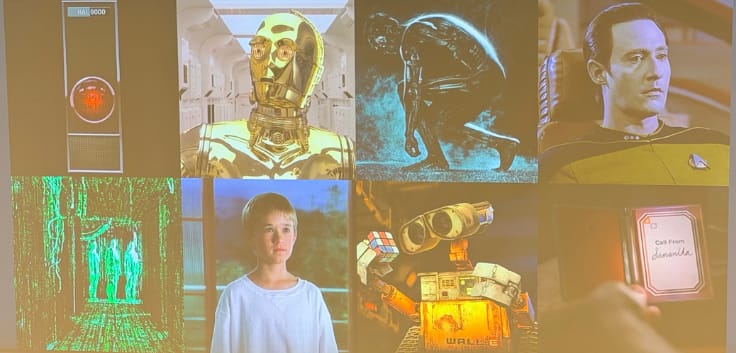 Alex Hartemink presenting from the front of a large lecture hall. His slide is titled "Humans have longed to make intelligent beings in their own image" and shows a collage of film stills