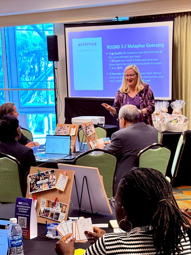 Woman presents at the front of the room. Behind her is a slide that reads: Metaphor guessing