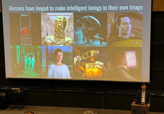 Alex Hartemink presenting from the front of a large lecture hall. His slide is titled "Humans have longed to make intelligent beings in their own image" and shows a collage of film stills, including HAL 9000, C-3PO from Star Wars, TRON, Data from Star Trek, a young boy in a white sweatshirt looking out a window toward mountains and trees, three shadowy green-iridescent figures in a room with glowing floors, walls, and ceiling, WALL-E holding a Rubik's cube, and a hand holding a foldable device showing a call from Samantha in the film Her.